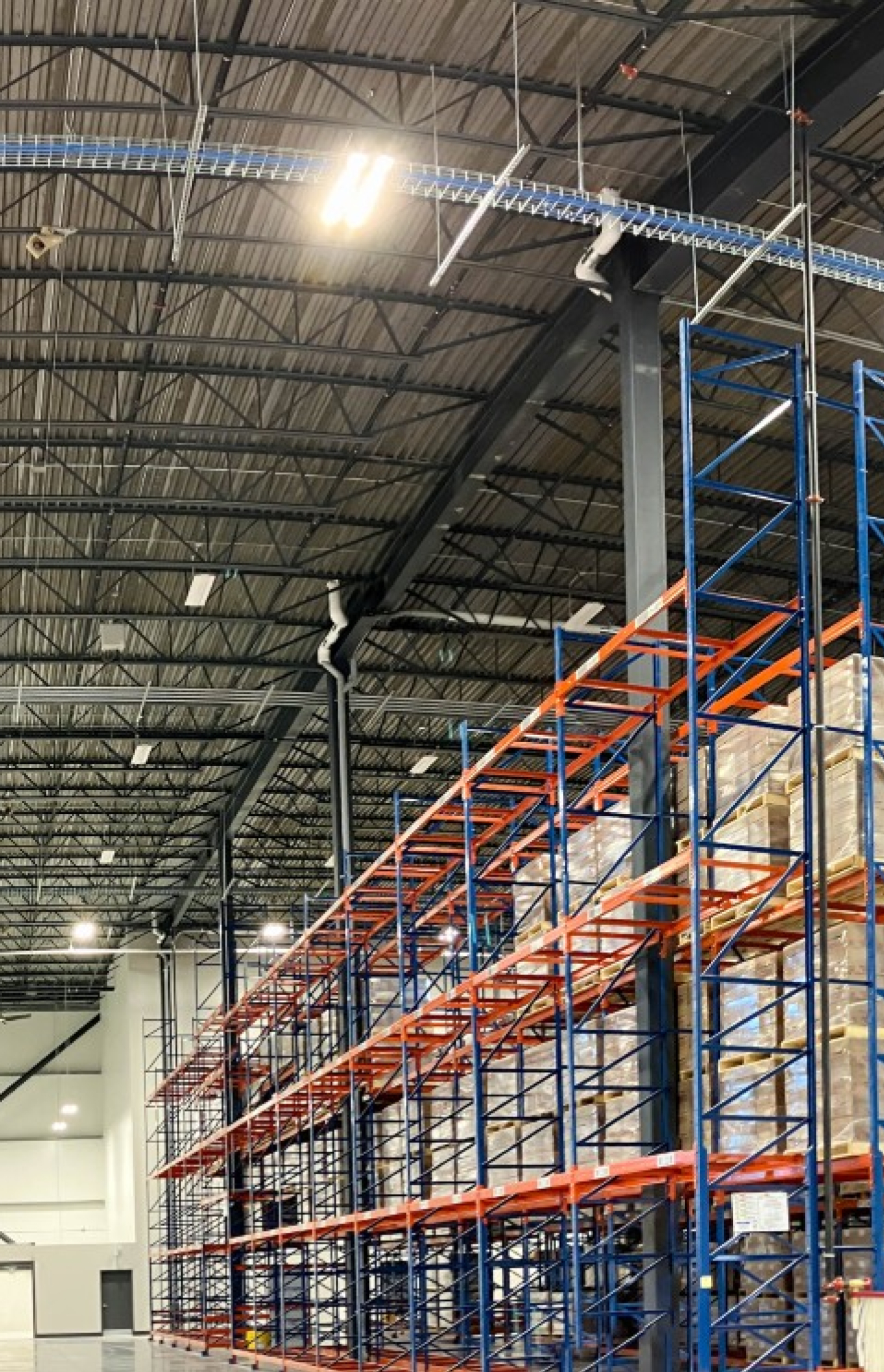 A view of a large warehouse interior with tall blue‑and‑orange storage racks filled with stacked pallets and boxes. Overhead lighting, steel roof structure, and equipment line the polished concrete floor.